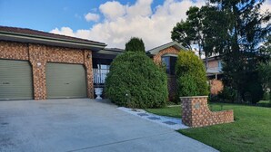 A residential driveway leading to a brick garage with a green door, a well-maintained lawn, and a stone planter with a small tree. - The Junction Estate. Cool, Calm and Cozy. (Wallerawang)