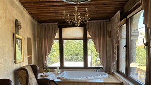 A room with a wooden ceiling, a large window with curtains, a bathtub, a dining table with chairs, and a chandelier. - Kadievi (Ürgüp)