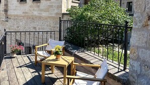 A wooden deck with outdoor furniture, a stone building with balconies, and a large tree. - Kadievi (Ürgüp)