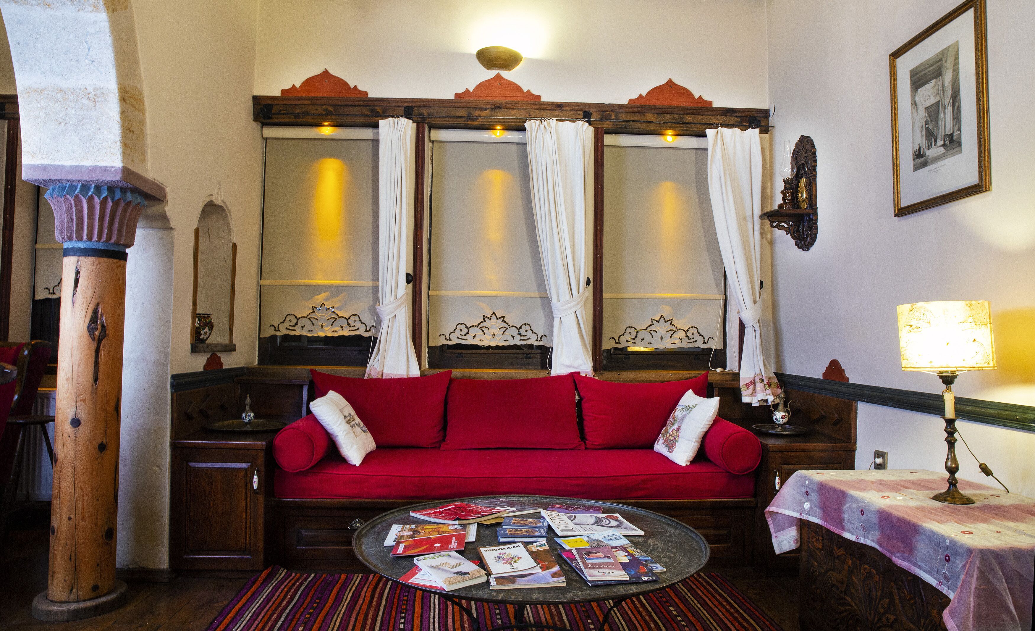 Un cuarto acogedor con un sofá rojo, una mesa redonda con revistas, una lámpara y un cuadro en la pared.