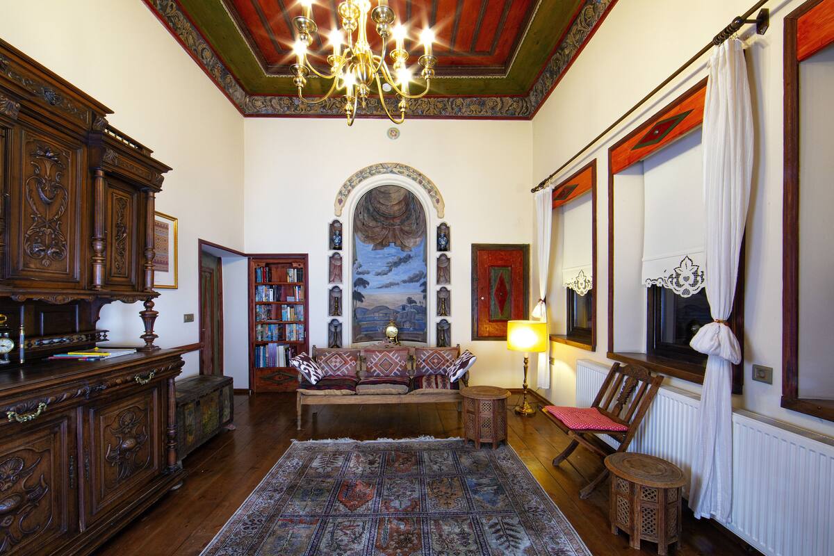 A traditional room with a wooden floor, a patterned rug, a sofa with cushions, a small round table, a bookshelf, and a chandelier.