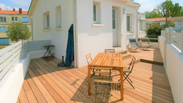 A white building with a wooden deck, a table with chairs, and a blue umbrella. - Charming Renovated House: Close to Beach & Town Center, Modern Amenities (La Tranche-sur-Mer)