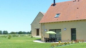Une maison à deux étages, avec un toit recouvert de tuiles rouges, une petite fenêtre à l’étage supérieur et une terrasse aménagée avec du mobilier d’extérieur.