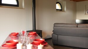 Un coin repas avec une longue table dressée pour quatre personnes, des bols rouges et des bougies blanches. On aperçoit une cheminée noire à l’arrière-plan.