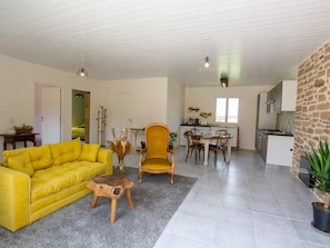 Ein Wohnzimmer mit gelber Couch, einem Holzstuhl und einem kleinen Tisch mit einer Vase. Es gibt eine Küchenzeile mit Esstisch und Stühlen.