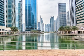 A waterfront view with modern high-rise buildings, a calm water body, and a paved walkway.