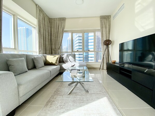 A modern living room with a grey sofa, a glass coffee table, a black TV stand, and large windows with curtains.