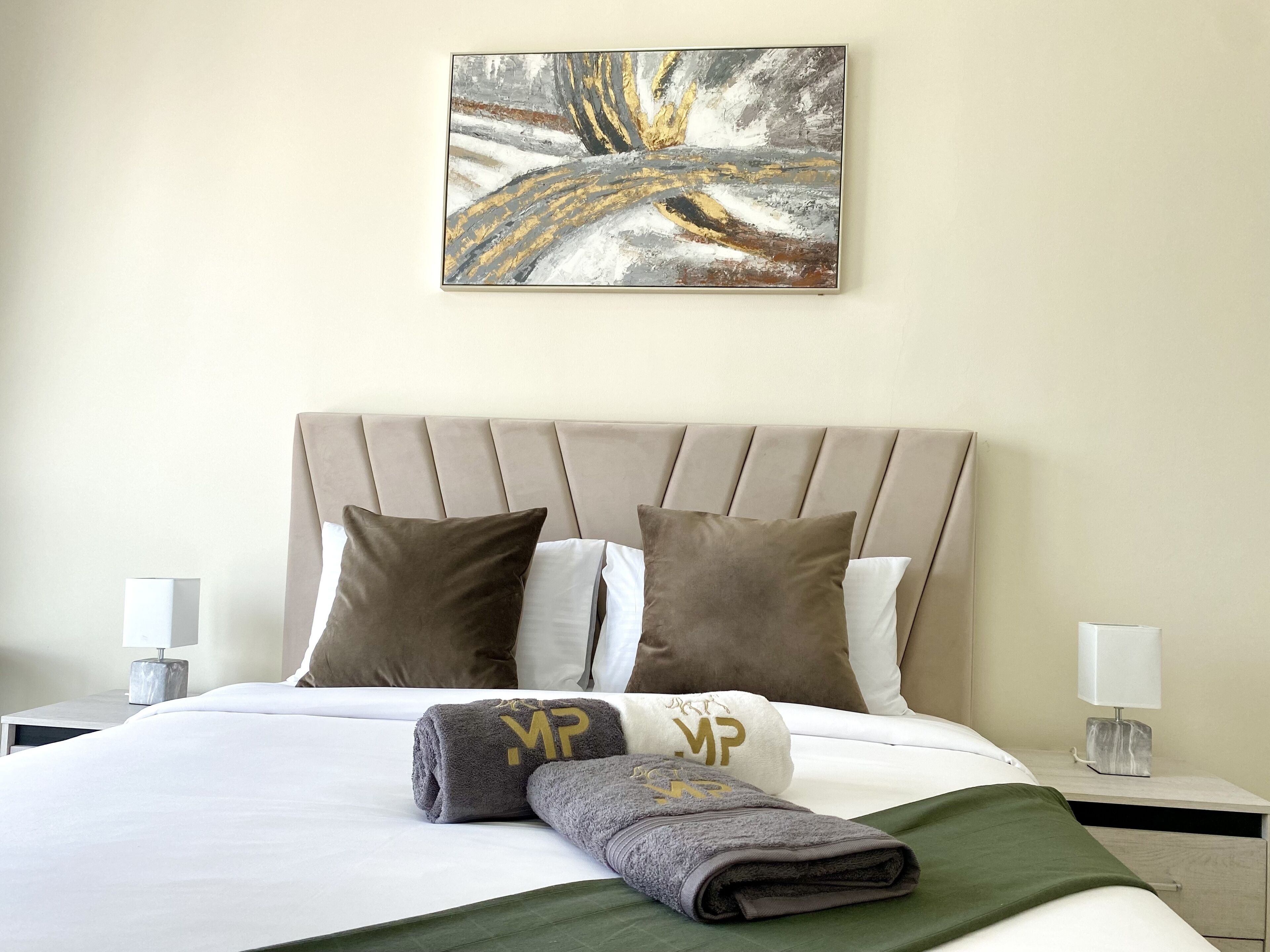 A neatly made bed with a beige headboard, two pillows, and a folded towel with a logo.