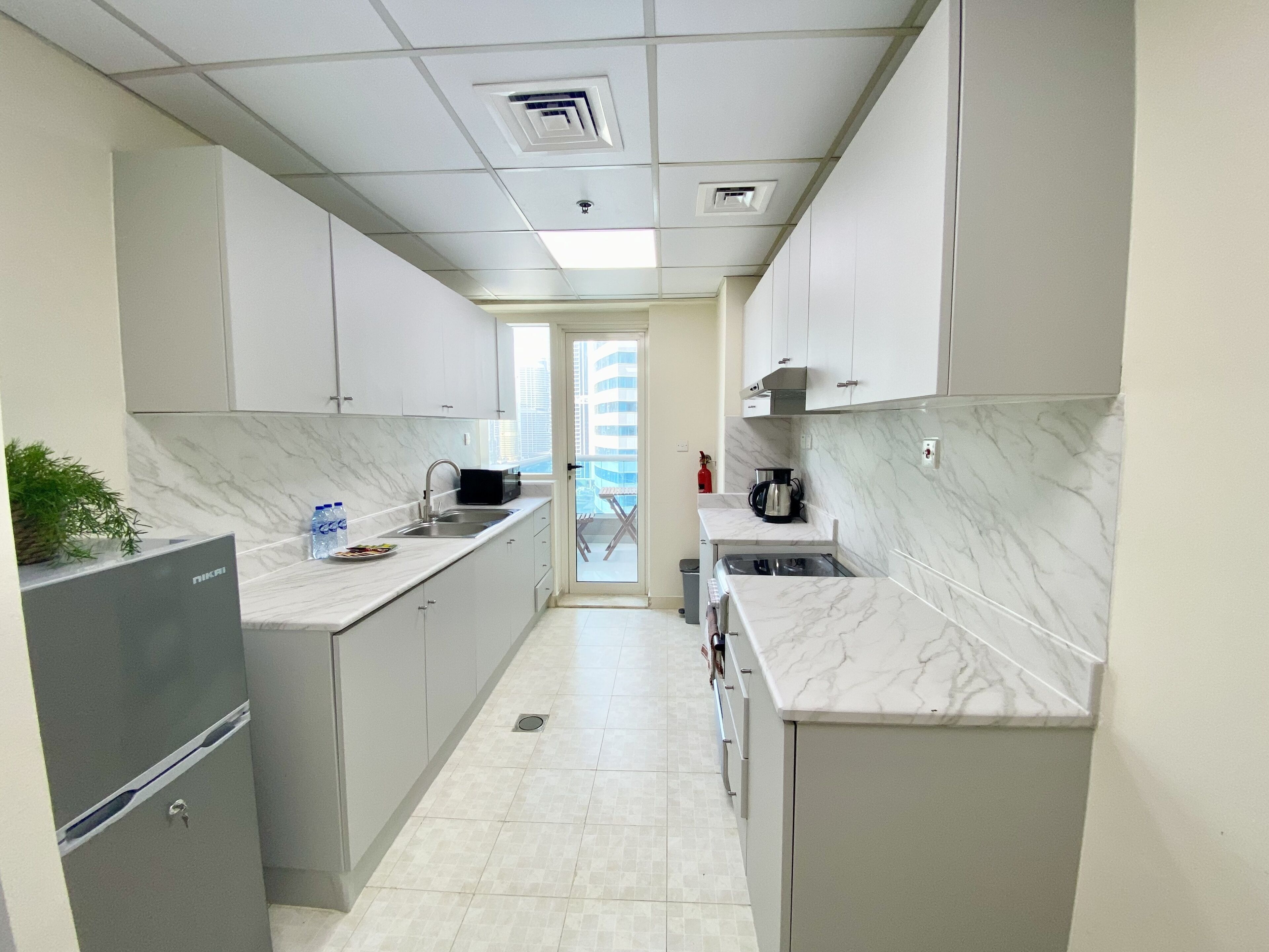 A narrow kitchen with white cabinets, a marble countertop, and a view of a balcony.