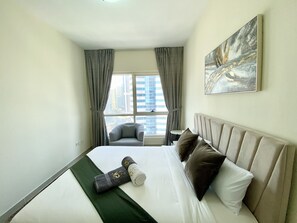 A hotel room with a bed, a chair, a painting, and a view of buildings.