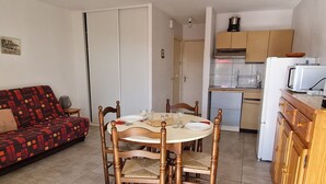 Un espace de vie compact comprenant une table à manger pour quatre personnes, entourée de chaises en bois. Un canapé rouge à motifs est placé contre le mur. Une petite cuisine équipée d’un four à micro-ondes et d’une cuisinière est visible sur la droite.