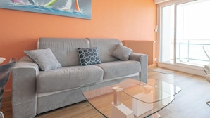 A modern living room with a grey sofa, a glass coffee table, and a large window with a view of the outdoors. - Modern 80m² apartment with wifi, dishwasher, balcony, and sea view (Les Sables-d'Olonne)