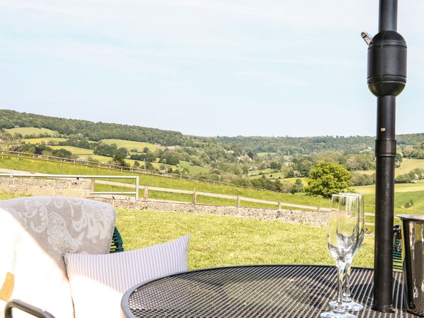 A table with a glass of water, a bottle, and a metal container, overlooking a grassy landscape with a wooden fence and hills in the distance. - Farm View Lodge (Stroud)