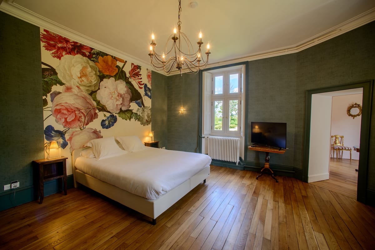 A bedroom with a large bed, a chandelier, a television, and a floral wall mural.