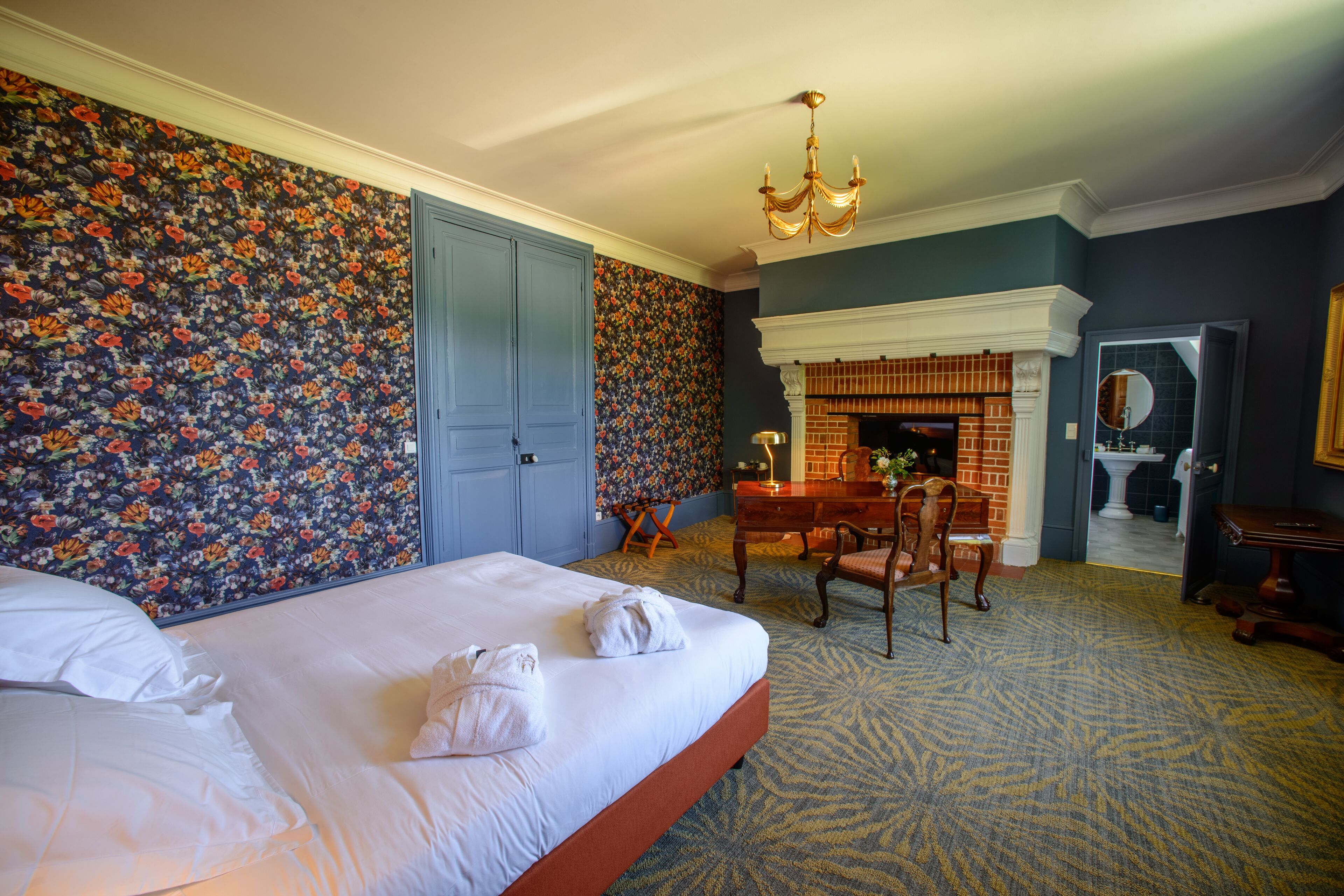 Habitación de hotel con una cama grande, un escritorio con una silla, una chimenea y un papel tapiz floral.