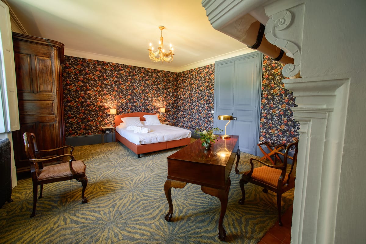 A bedroom with a bed, two chairs, a table with a vase, a chandelier, and a door.