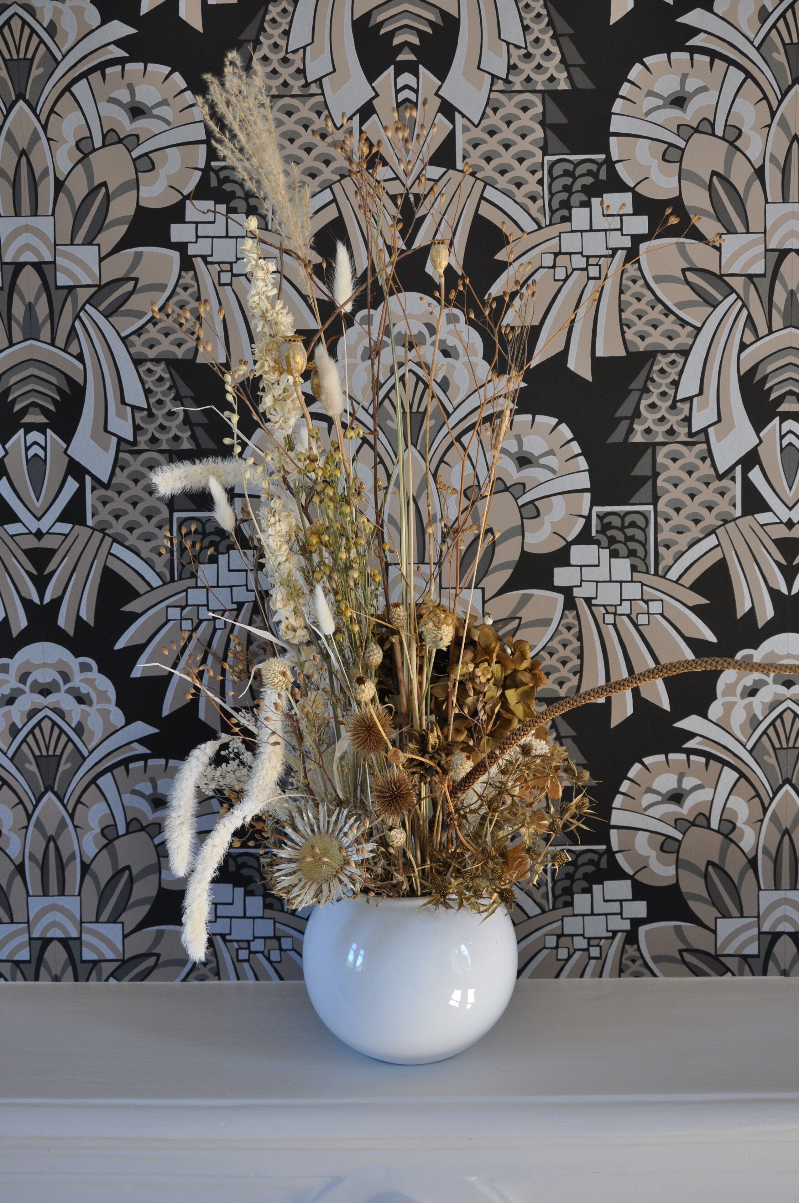 A white vase with dried flowers and pampas grass on a white surface against a patterned wallpaper background.