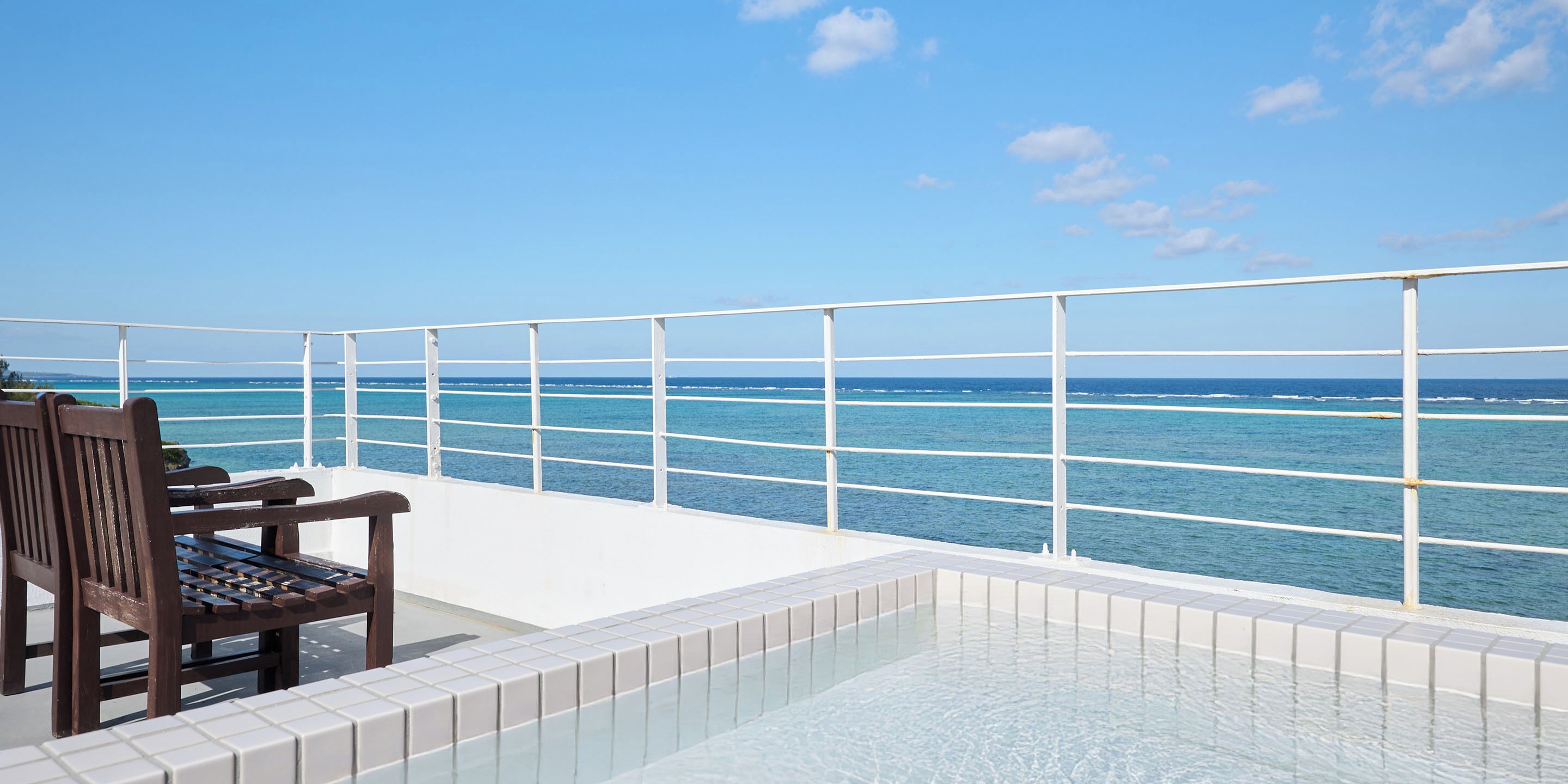 A pool area with a wooden bench and a white railing overlooking the sea.