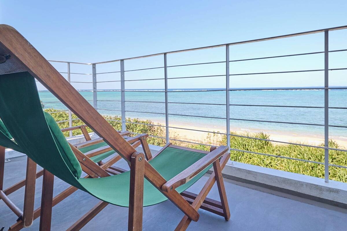 Ein Hängestuhl aus Holz mit einem grünen Kissen auf einem Balkon mit Blick auf einen Strand.