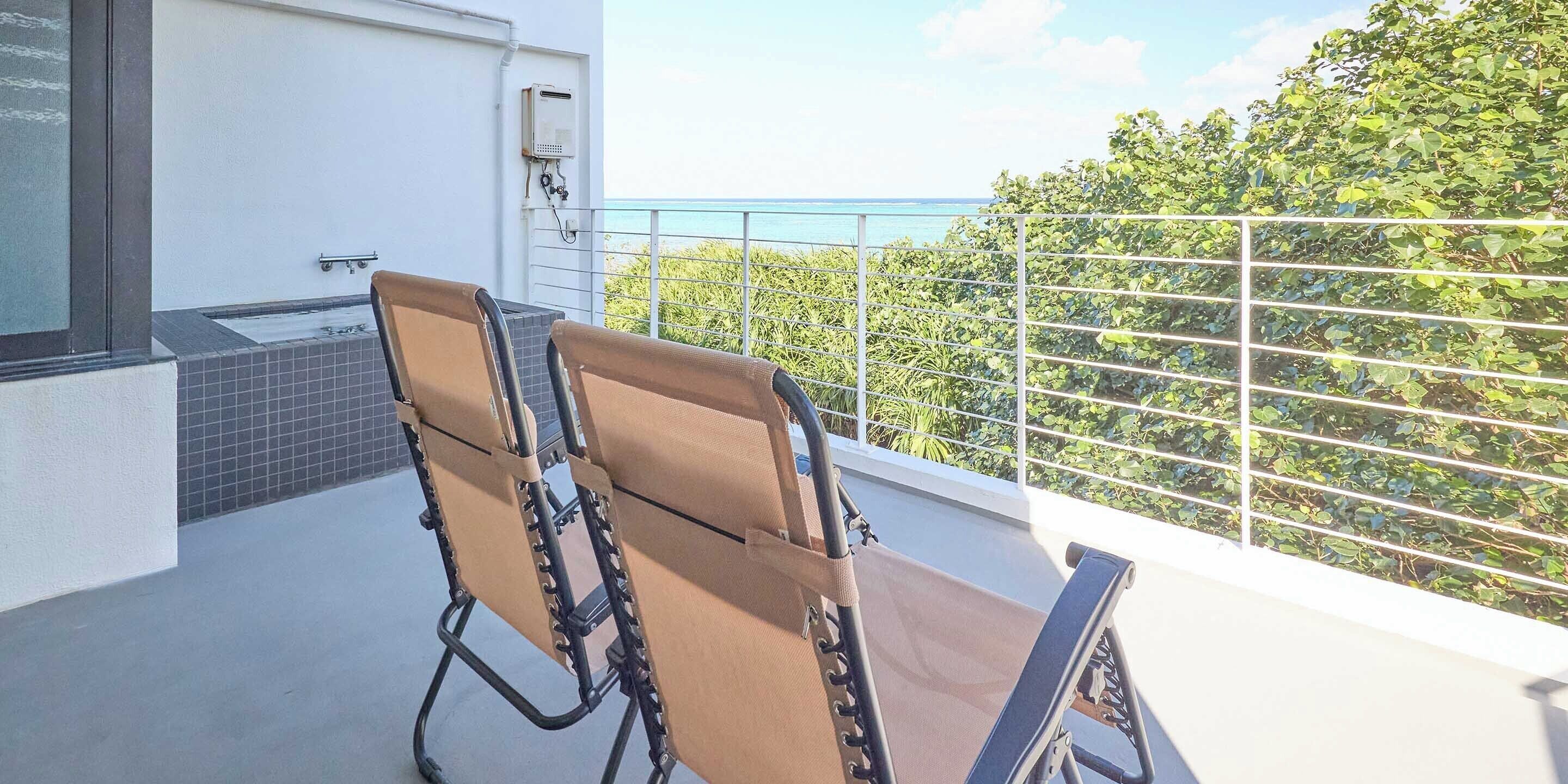 A balcony with two sun loungers, a white wall, a tiled area, and a view of the sea and greenery.