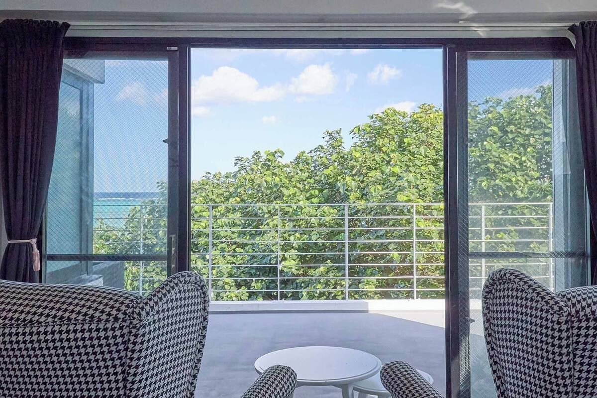 Ein Balkon mit zwei Chesterfield-Sofas mit Hunde-Zahn-Muster, einem kleinen runden Tisch und Blick auf eine grüne Baumlinie durch ein Metallgitter.