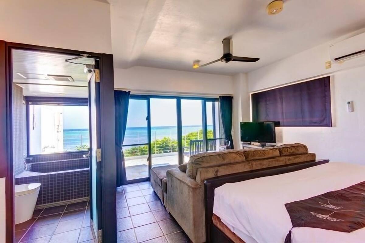 Ein Hotelzimmer mit Bett, Fernseher, einem Balkon mit Meerblick und einer Duschkabine.