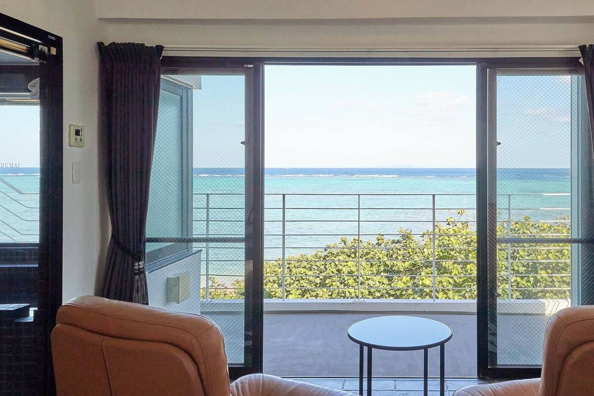Ein Zimmer mit einer großen Schiebeglasdoor zum Balkon mit Meerblick. Auf dem Balkon befinden sich zwei bequeme Stühle und ein kleiner Tisch.