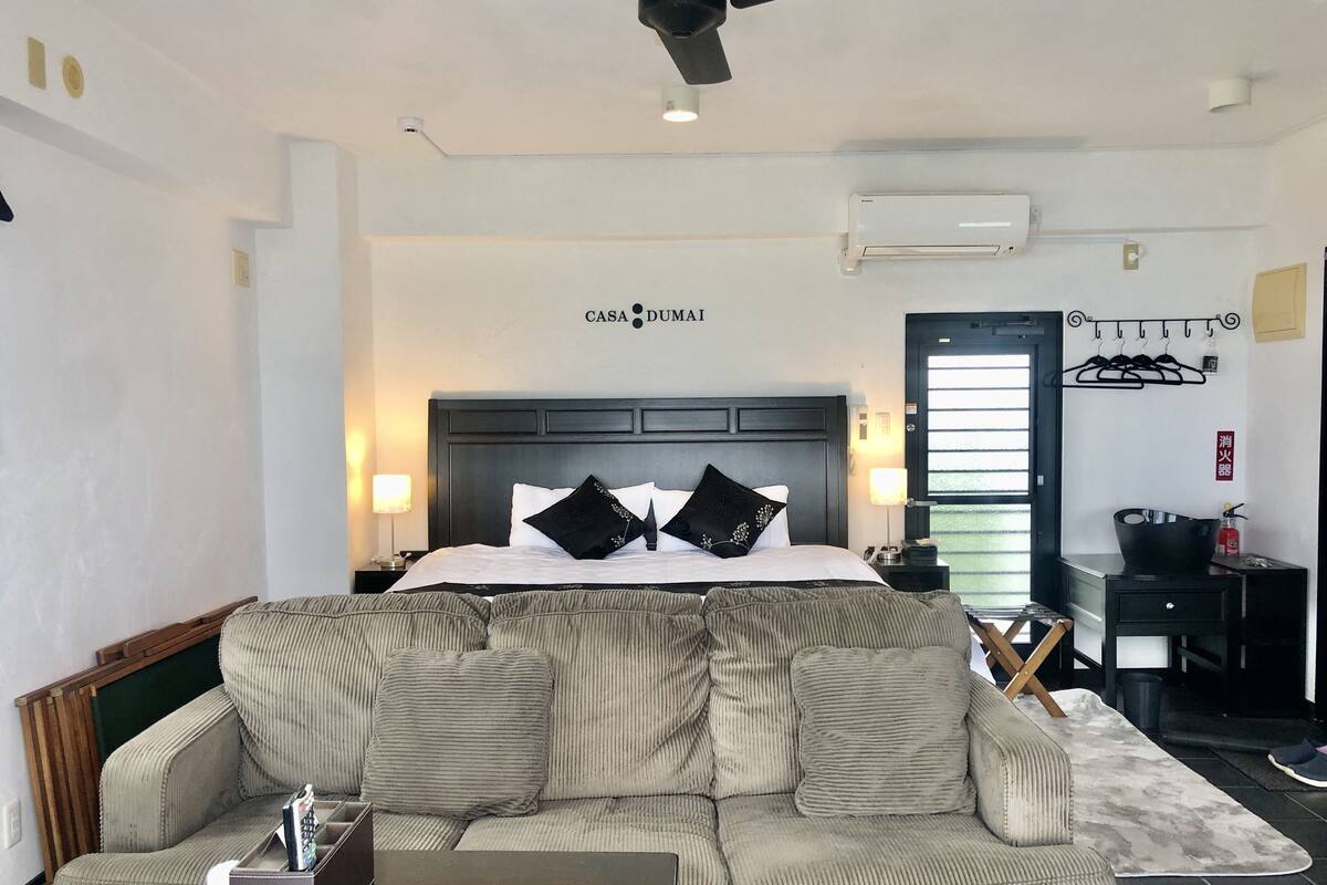 Ein Hotelzimmer mit einem Bett, einer Couch, einem Deckenventilator, einer Klimaanlage, einer Wandleuchte und einer Tür mit Griff.