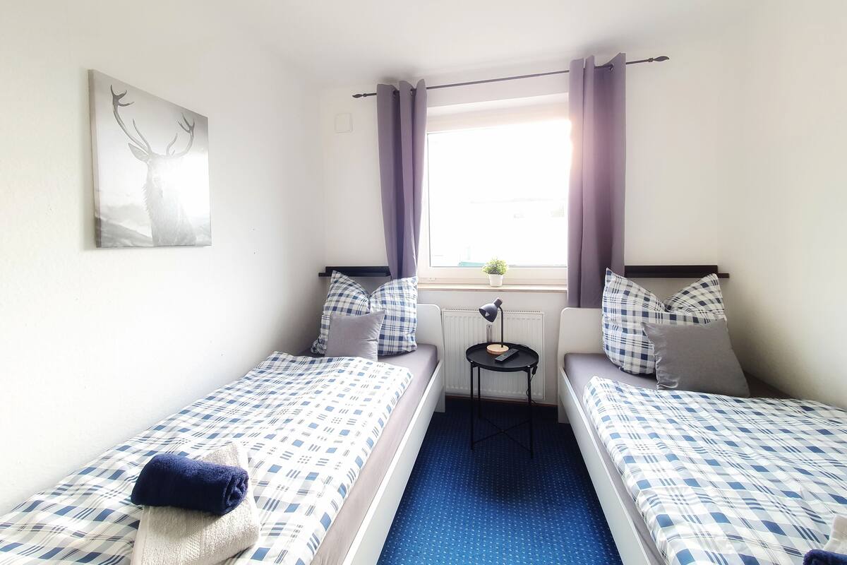 Una habitación con dos camas, una mesita, una ventana con cortinas e imagen colgada en la pared.