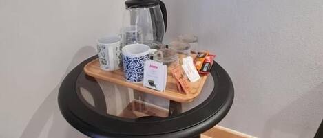 A small round table with a glass top, a coffee maker, a mug, a cup, a box, and some snacks.