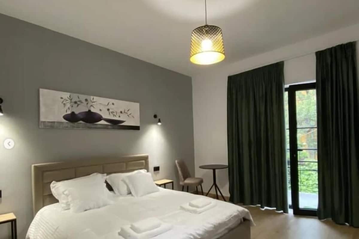 A modern bedroom with a large bed, a painting on the wall, a small table, and a view of greenery through the window.