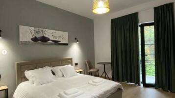 Un dormitorio moderno con una cama grande, un cuadro en la pared, una mesita pequeña y vistas a zonas verdes a través de la ventana.