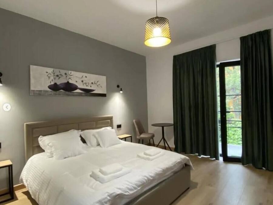 Un dormitorio moderno con una cama grande, un cuadro en la pared, una mesita pequeña y vistas a zonas verdes a través de la ventana.