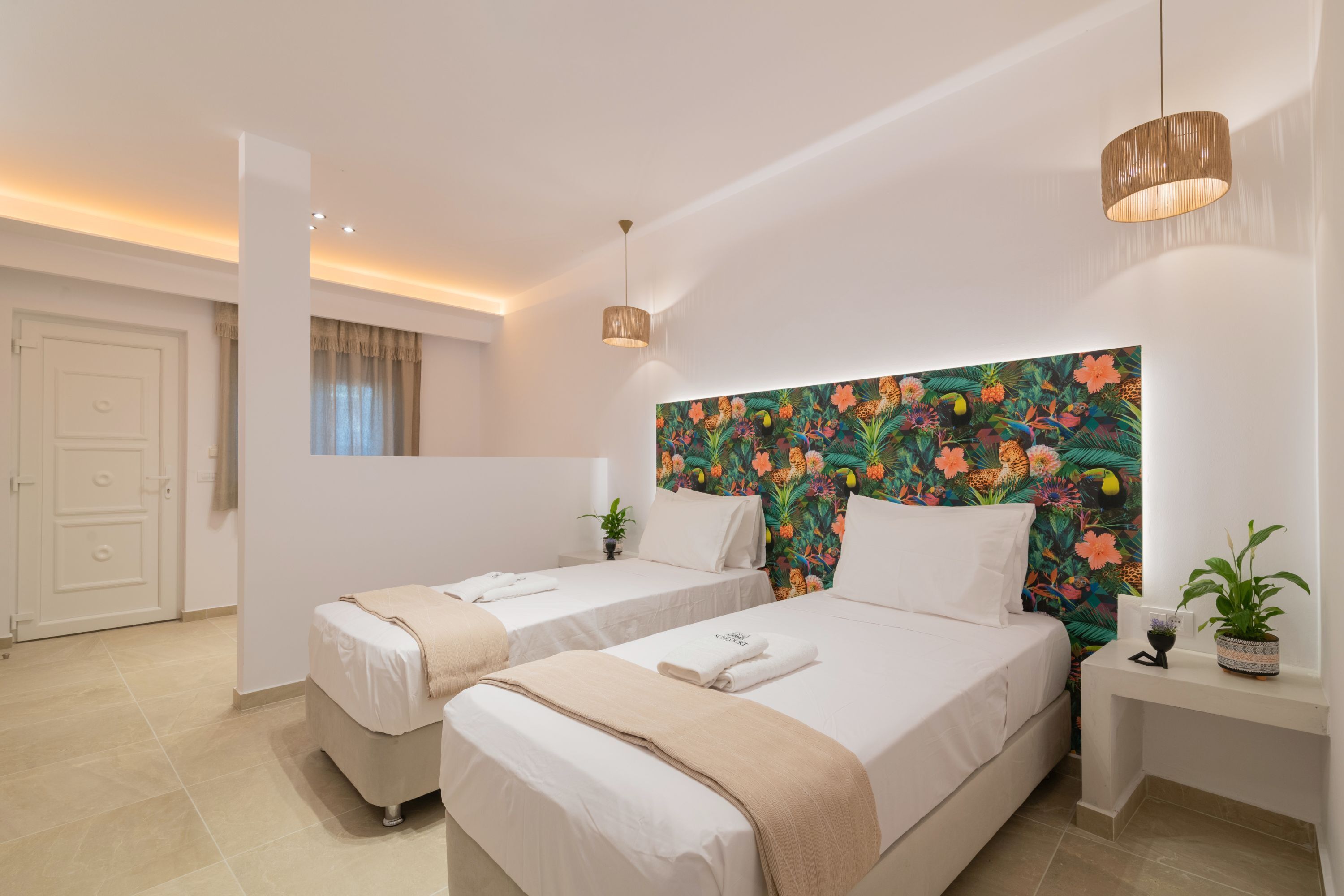 A hotel room with two beds, a colorful floral headboard, a small bedside table with a plant, and a door with a curtain.
