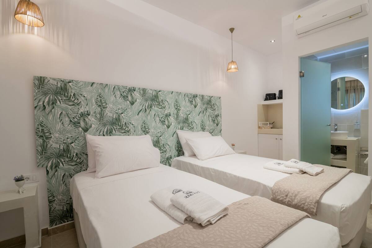 Hotelkamer met twee bedden, een groot hoofdeinde met een bladerenpatroon, een nachtkastje met een lamp, en een badkamer met spiegel en wastafel.