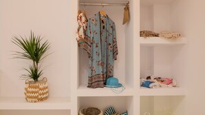 Une pièce bien rangée, avec une plante en pot, des vêtements suspendus, un panier et une étagère où sont pliés des couvertures et un chapeau.