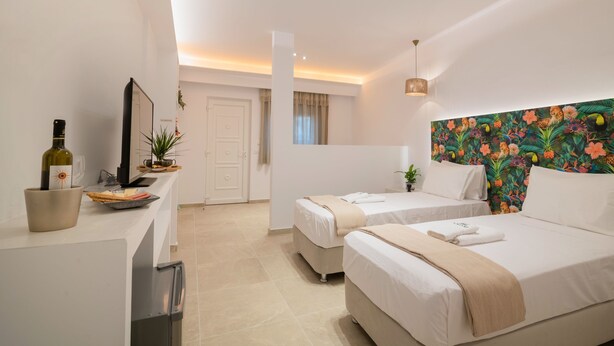 Une chambre d’hôtel avec deux lits, une kitchenette, une grande fenêtre et une tête de lit ornée de motifs floraux vifs.