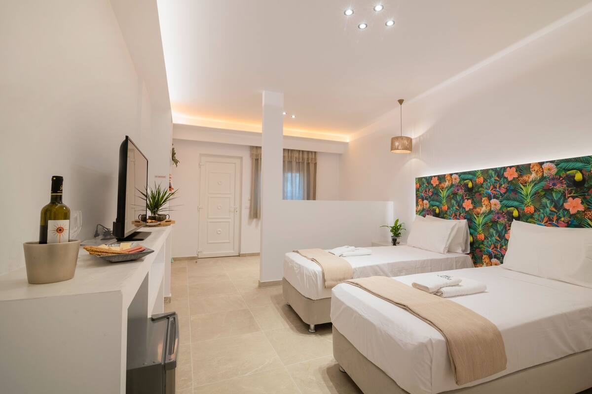 Een hotelkamer met twee bedden, een kitchenette, een groot raam en een kleurrijk hoofdeinde met bloemen.