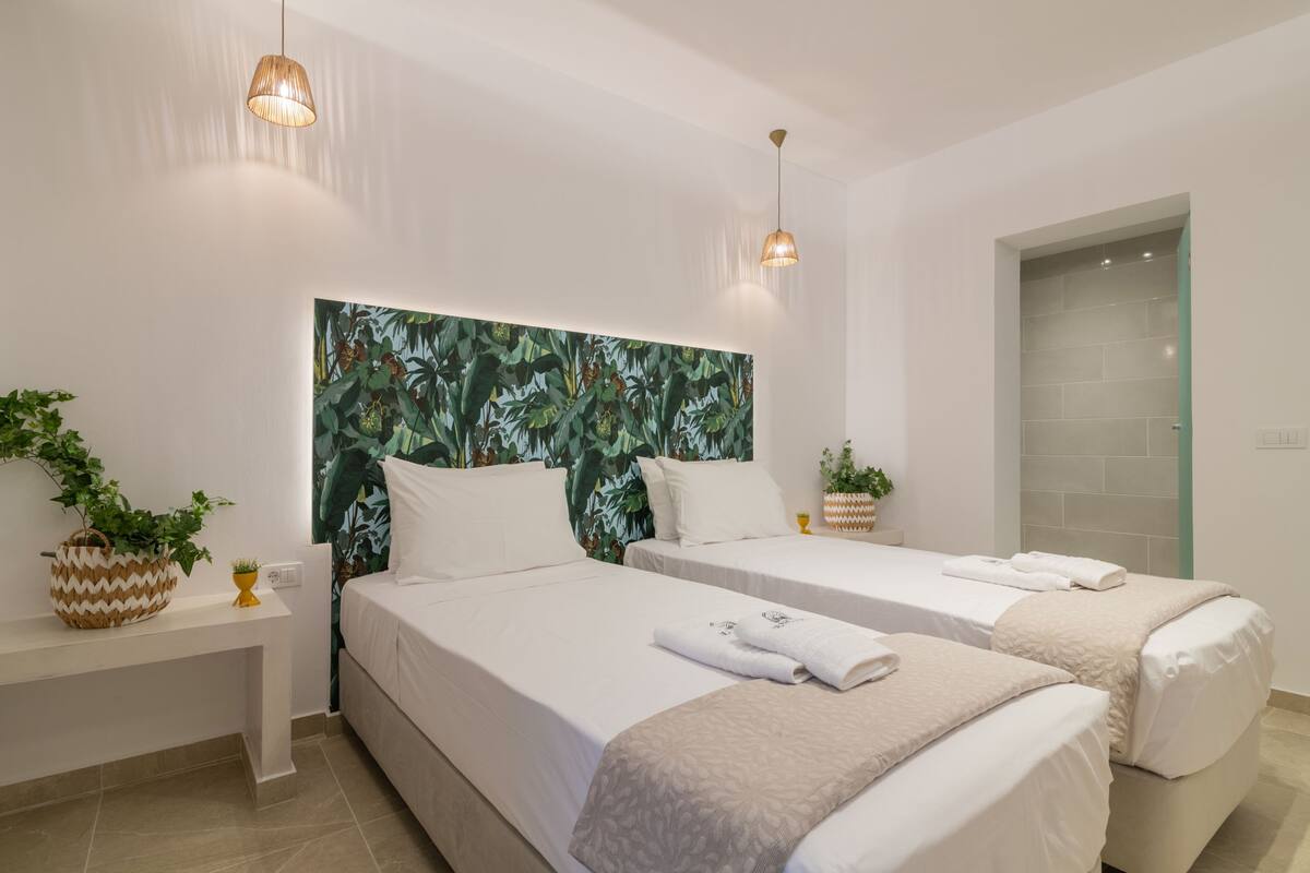 Hotelkamer met twee bedden, een groot hoofdbord met tropisch thema, nachtkastjes met potplanten en hanglampen.