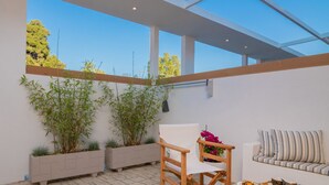 Une terrasse moderne avec un canapé blanc, une chaise en bois et une table où sont disposés des aliments. On y trouve deux jardinières avec des plantes vertes et une vue dégagée sur le ciel.