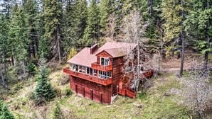 A two-story wooden cabin with a balcony, surrounded by a forested area.