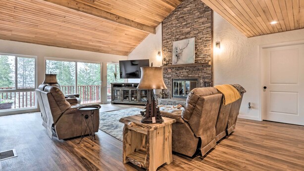 A spacious living room with a stone fireplace, wooden ceiling, and a large window overlooking a deck.