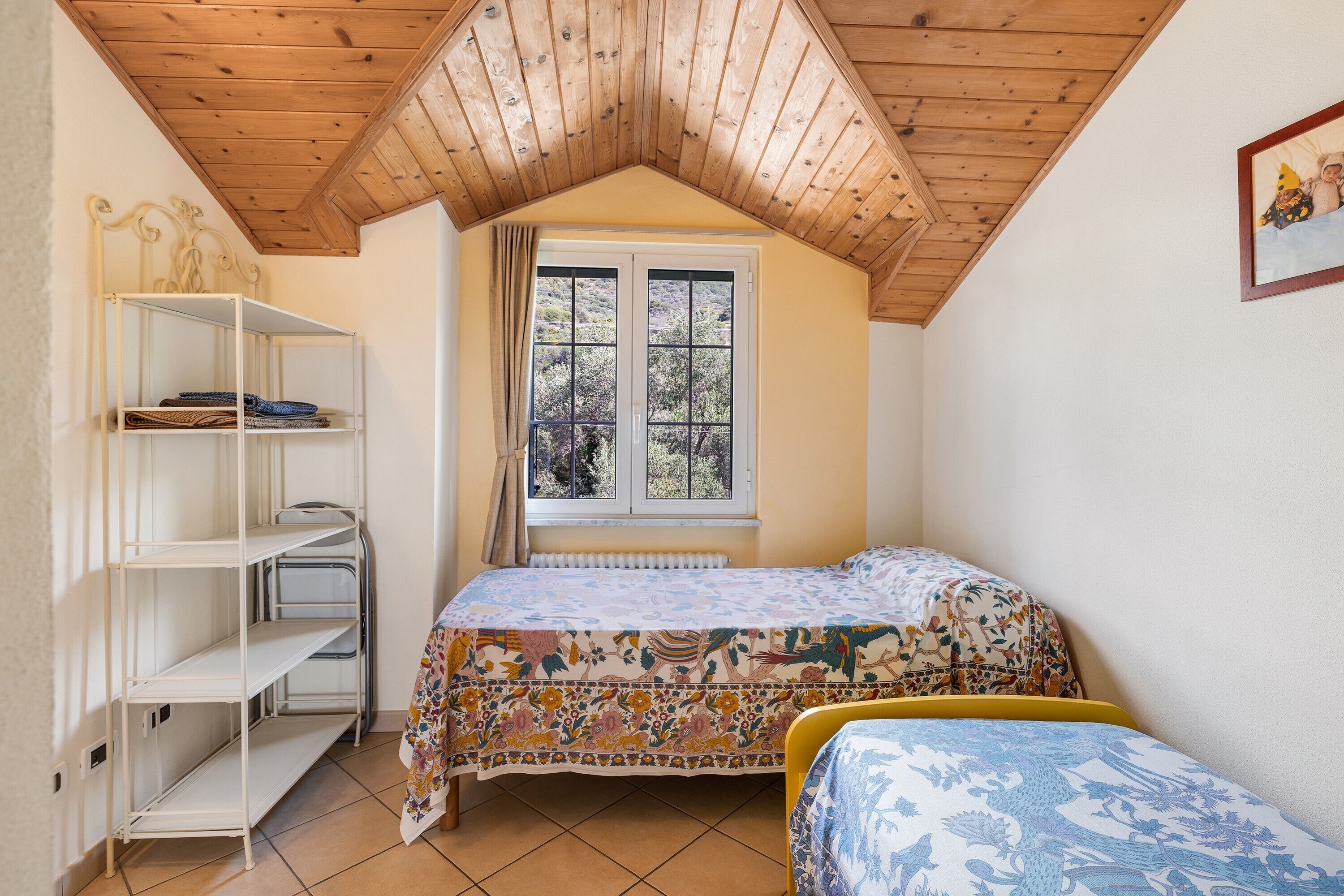 Ein Schlafzimmer mit schrägen Wänden, einem Fenster mit Ausblick, einem Bett mit Blumenmuster-Decke und einer weißen Regaleinrichtung.