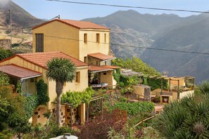 Une maison située sur une colline, avec un toit recouvert de tuiles rouges, un petit balcon et un jardin où poussent différentes espèces de plantes.