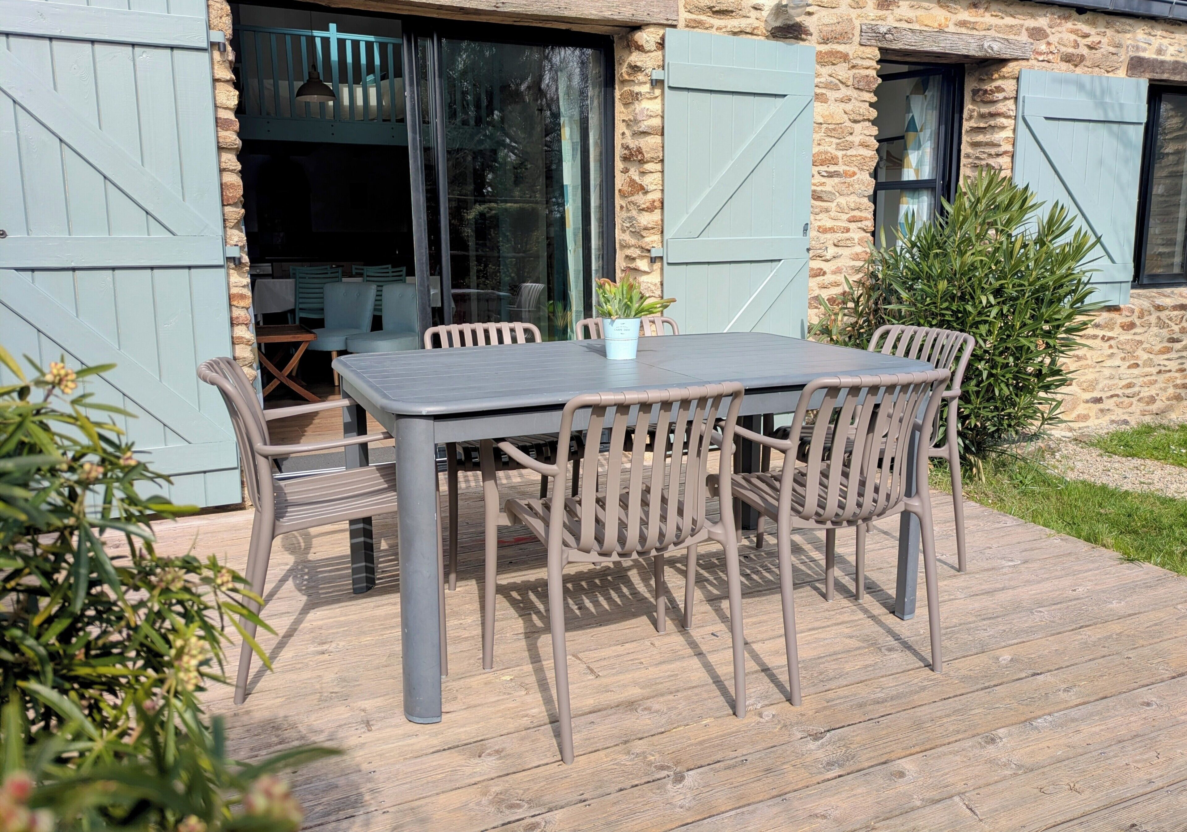 Une terrasse avec une table et des chaises, un pot de fleurs et un bâtiment en pierre avec des volets.
