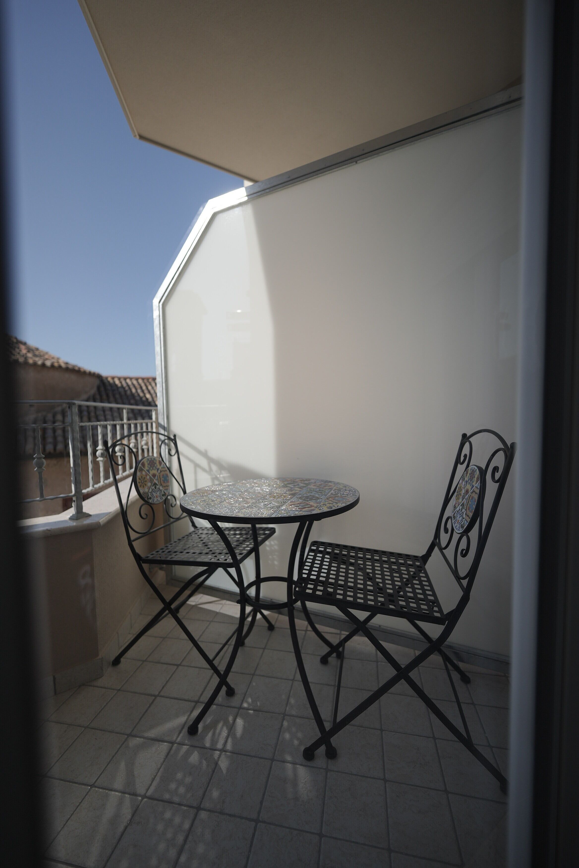 Un balcón con una pequeña mesa, dos sillas, una ventana y piso de baldosas.