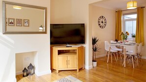 A living room with a flat-screen TV, wooden cabinet, dining table with chairs, a clock, and a window with curtains. - 2BR Stylish Norwich Stay Near Cathedral | Parking (Norwich)