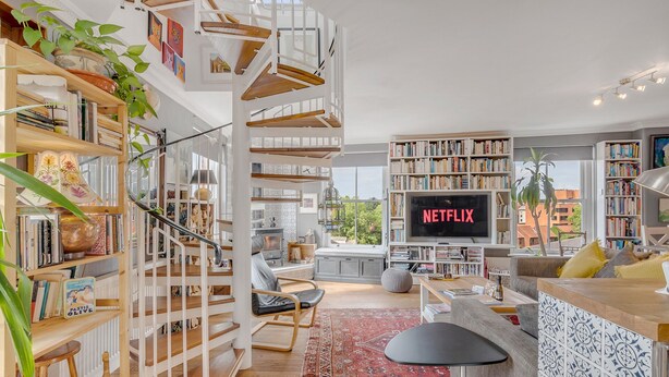 Amplia sala de estar con techo alto, una escalera central y una gran estantería llena de libros. Un televisor de pantalla plana está montado en la pared, y hay una zona de estar cómoda con un tapete rojo.