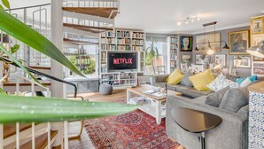 Un salón amplio con escalera, una gran estantería, un televisor mostrando 'NETFLIX' y un rincón de descanso acogedor.