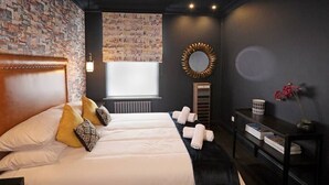 Et moderne hotelværelse med en væg i murstensmønster, en stor seng med hvide sengetøj, et rundt spejl og et lille bord med bøger og en vase.